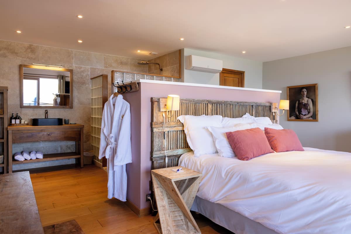 A bedroom with a large bed, a wooden bedside table, a nightstand, a mirror, a robe hanging, and a bathroom with a sink and shelves.
