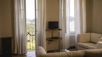 Un soggiorno con un divano, una televisione su un mobile basso e vista su un lago attraverso ampie finestre con tende leggere.