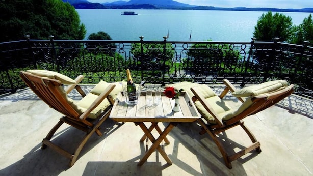 Un balcón con dos sillones de madera y una pequeña mesa, con vista a un lago que incluye un bote y montañas al fondo.