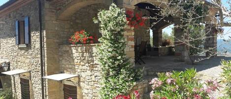 A stone house with a covered porch, a small garden with red flowers, and a view of the sea.