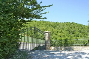 Una entrada con reja, un pilar de piedra y abundante vegetación, rodeada de colinas.