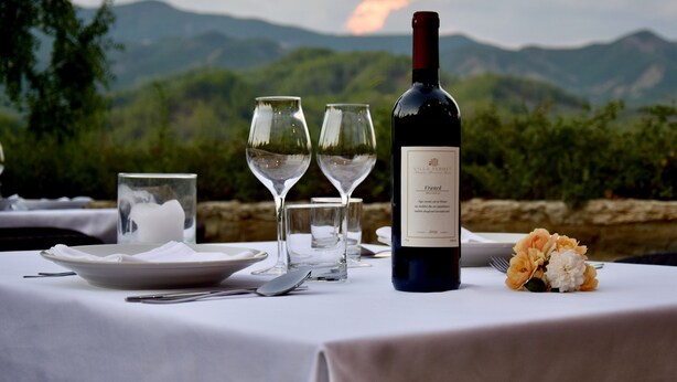 Una mesa con una botella de vino, copas y un plato, con vistas a un paisaje montañoso.