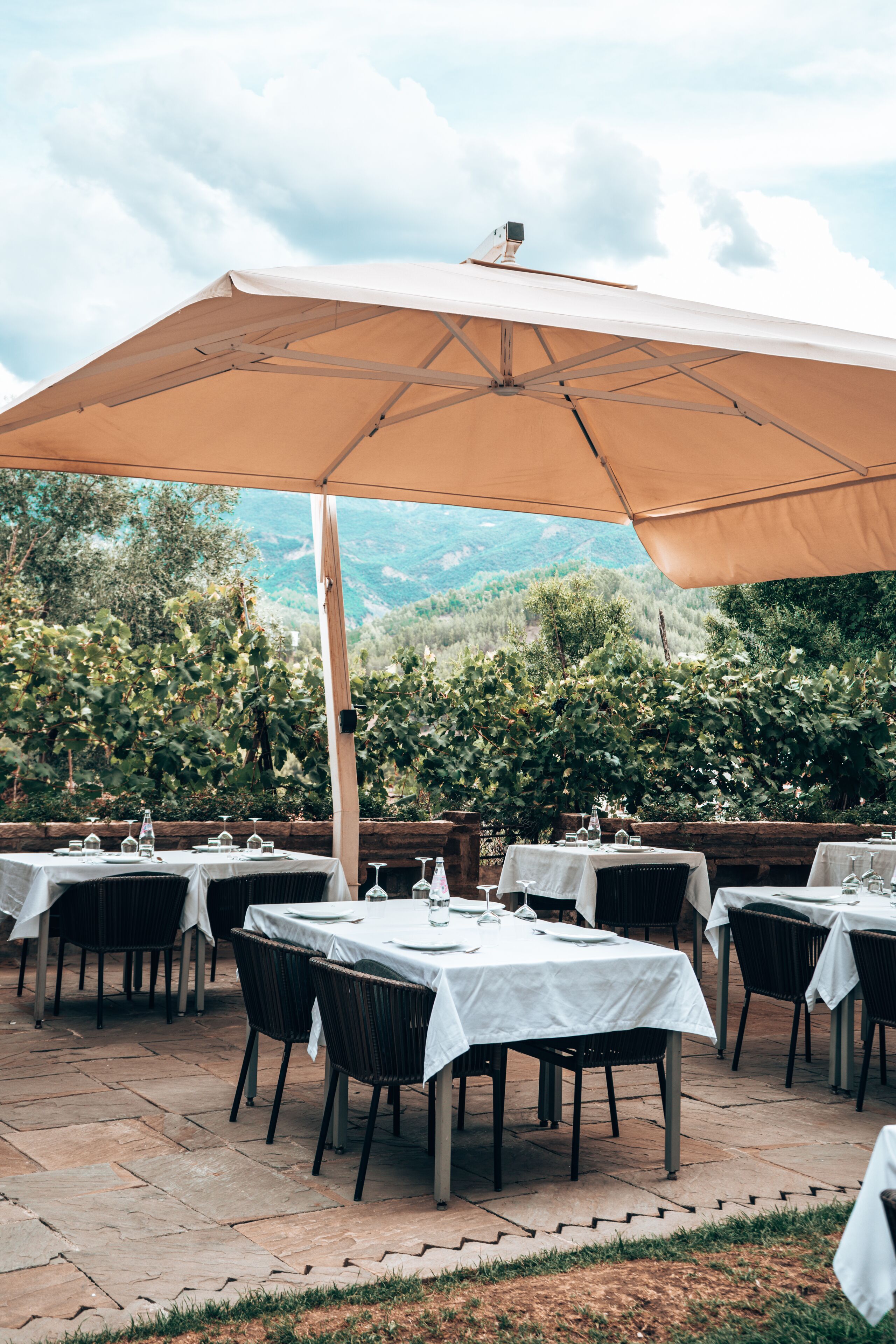 Een terras met tafels en stoelen onder een grote parasol, met een achtergrond van groen en heuvels.