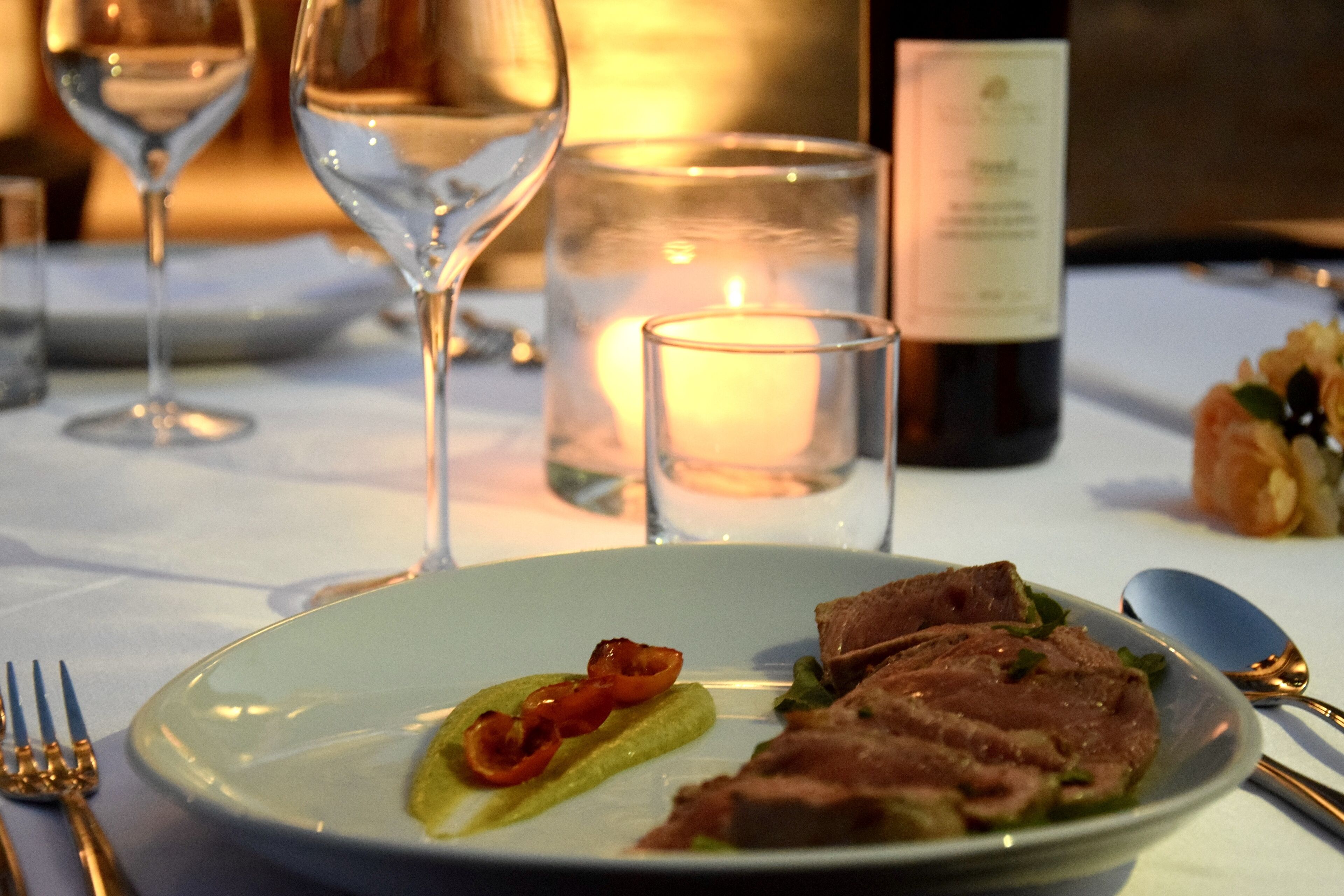A dining table with a plate of food, a wine glass, a candle, and a bottle of wine.