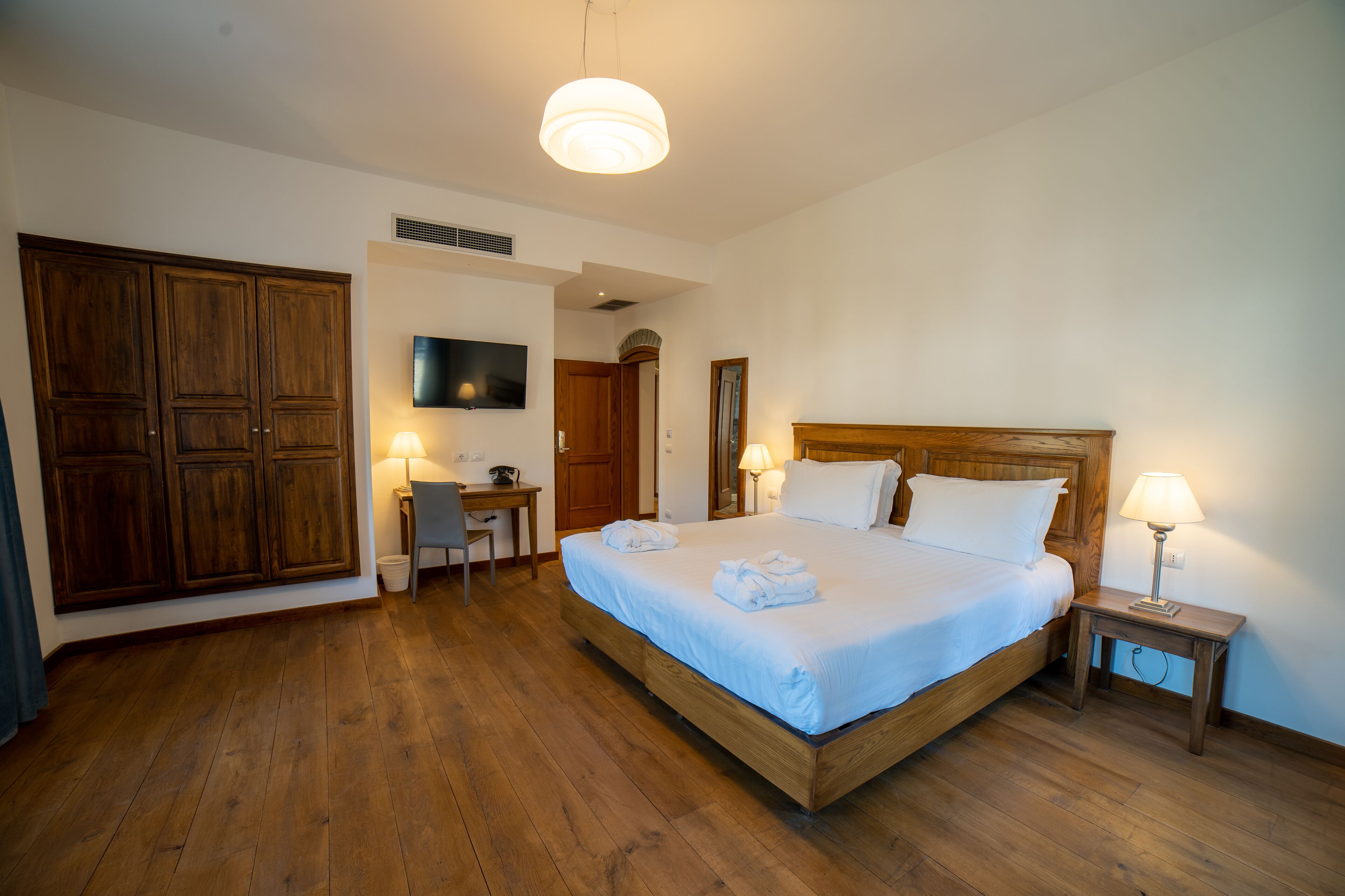 A bedroom with a wooden bed, two bedside lamps, a desk with a chair, and a TV mounted on the wall.