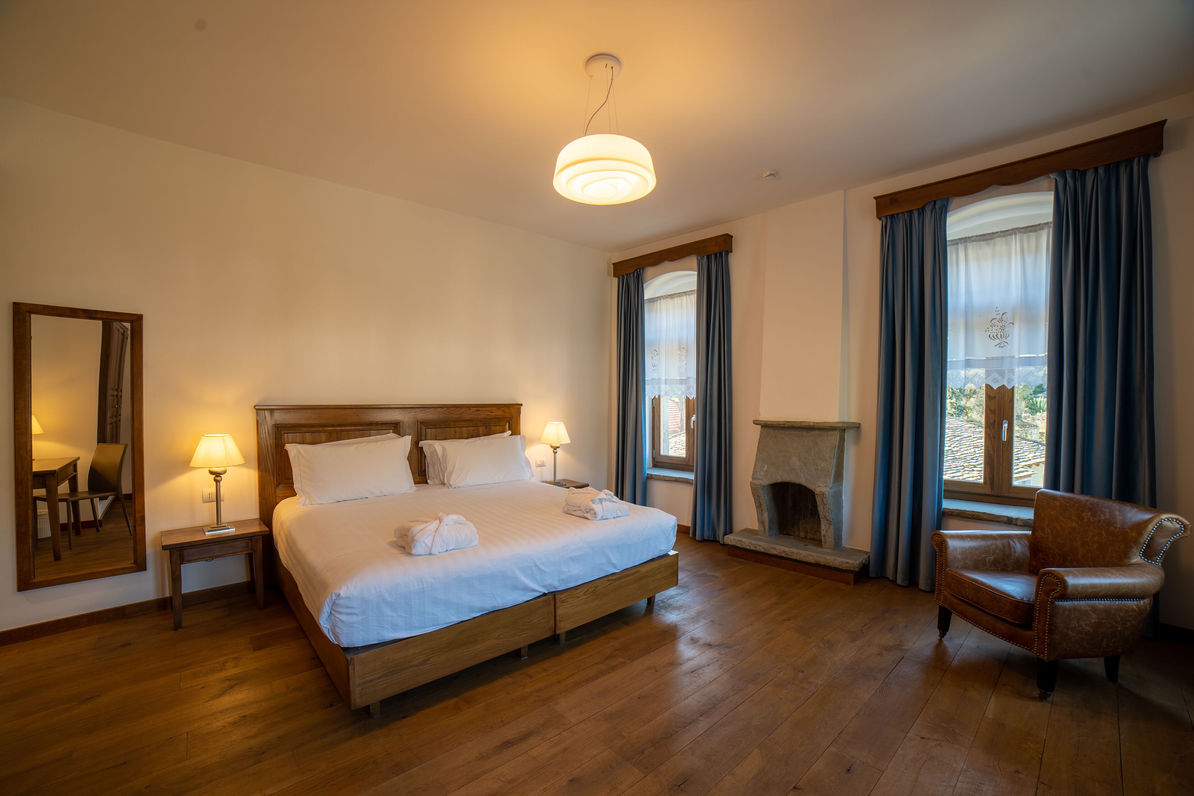 Una camera d'albergo con un letto di legno, lampade sui comodini, una poltrona, un camino e un'ampia finestra con tende.