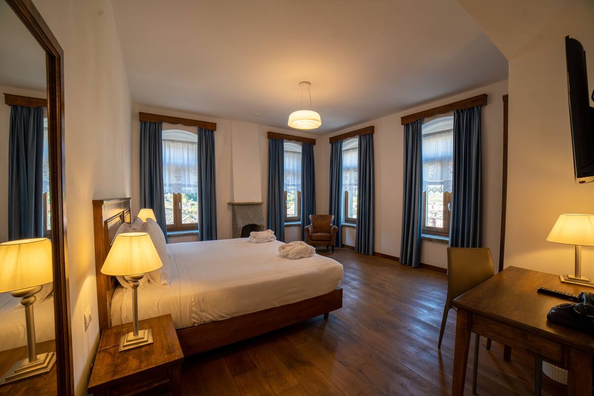 Habitación de hotel con cama, dos mesitas de noche, una silla, escritorio, televisor y ventanas grandes con cortinas.