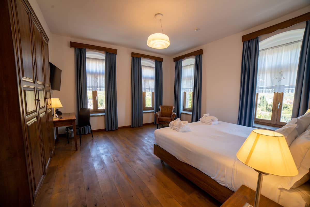 Habitación de hotel con piso de madera, una cama grande, un escritorio con lámpara, una silla y un televisor en la pared.