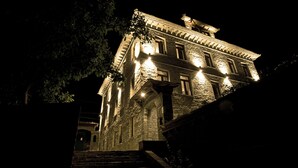 Un edificio de piedra con múltiples ventanas iluminadas y una entrada imponente, que sugiere una propiedad histórica o de lujo.