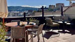 A rooftop terrace with a round wooden table, white and green chairs, and a view of a cityscape and mountains in the background. - Spectacular penthouse with terrace (Barcelona)