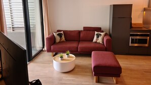 A modern living room with a red sofa, a round coffee table, and a small ottoman. There is a kitchen area with a built-in oven and a dining table. - Magnificent T2 parahotelier with 23m2 south-facing terrace and sea view (Ajaccio)