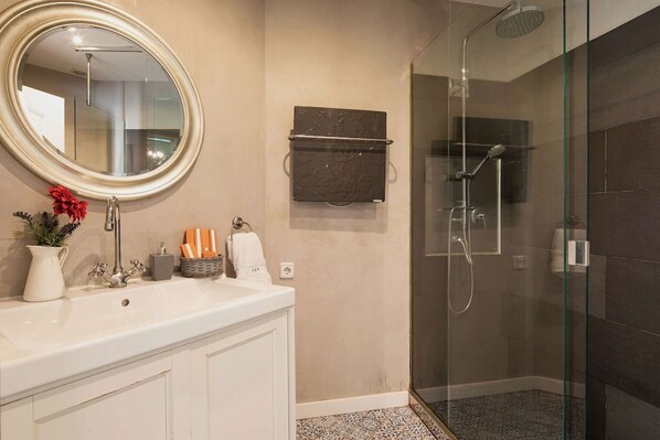 A bathroom with a round mirror, a white sink, a glass shower enclosure, and a dark tiled floor. - Bright modernist apartment in Rambla Catalunya (Barcelona)