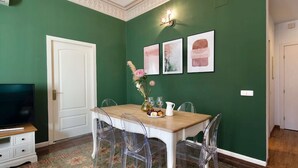 A dining area with a green wall, a white door, and a table with clear chairs. There are three framed pictures on the wall, a vase with flowers, and a small cabinet with a television. - Bright modernist apartment in Rambla Catalunya (Barcelona)