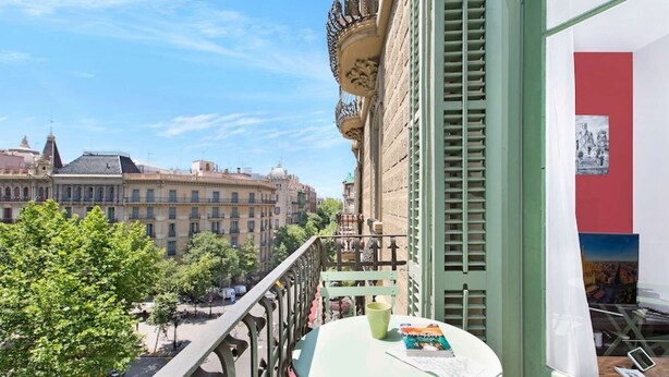 Un balcón con una mesa, barandilla verde y vista de edificios al fondo.