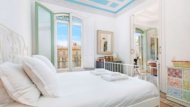 A bedroom with a bed, white bedding, a ceiling with blue accents, and a view of buildings through open doors. - Luxury and design in Rambla Catalunya (Barcelona)