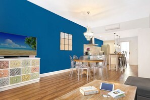 A modern living room with a blue accent wall, a wooden coffee table with books and a tablet, a dining area with clear chairs, and a kitchen in the background.