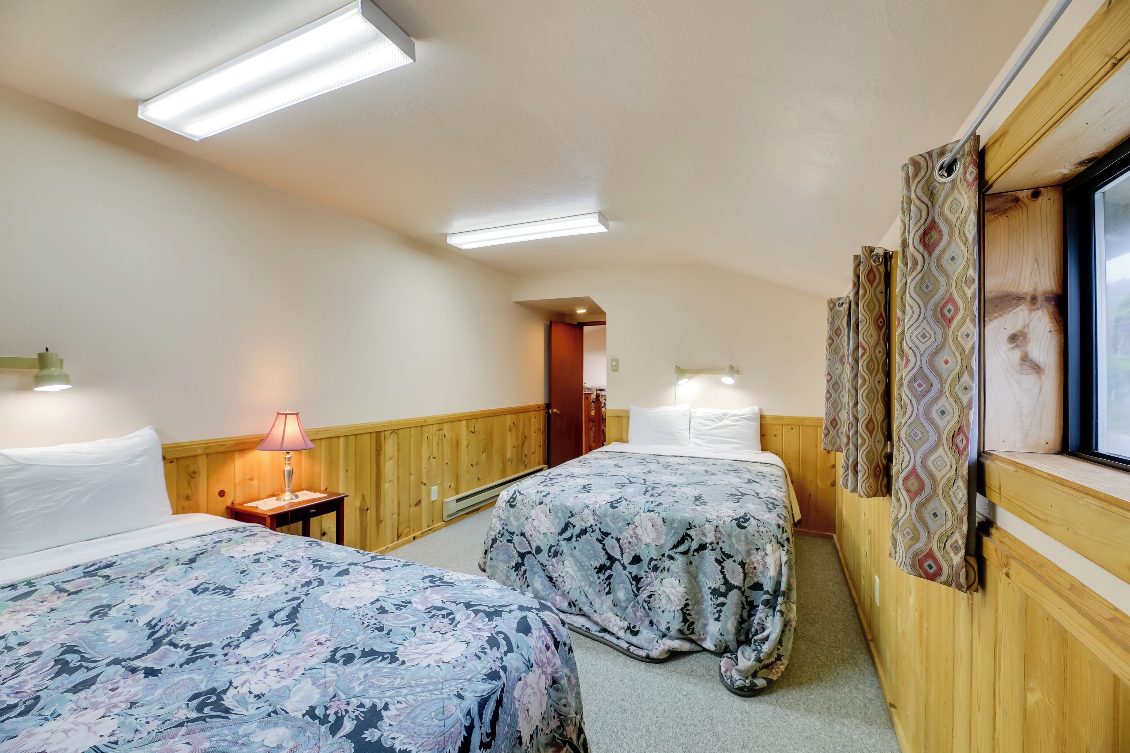 Ein Zimmer mit zwei Betten, Holzwänden, einem Fenster mit Vorhängen und einer Lampe.