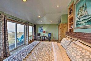 A hotel room with a large bed, a desk with two chairs, a painting, and a view through the window. - Sanderling Sea Cottages, Unit 9 with Ocean Views! (Waldport)