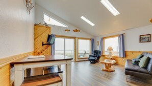 A spacious living area with a wooden floor, a sofa, a chair, a table, and a TV mounted on the wall.