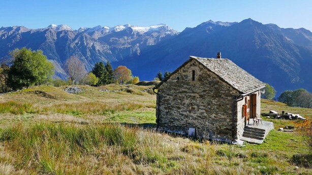 Une petite maison en pierre avec une porte en bois et des marches, entourée de pelouse et d’arbres, avec une chaîne de montagnes en arrière-plan.