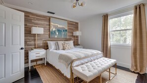 A bedroom with a bed, bedside tables, a bench, a painting, and a window with curtains.