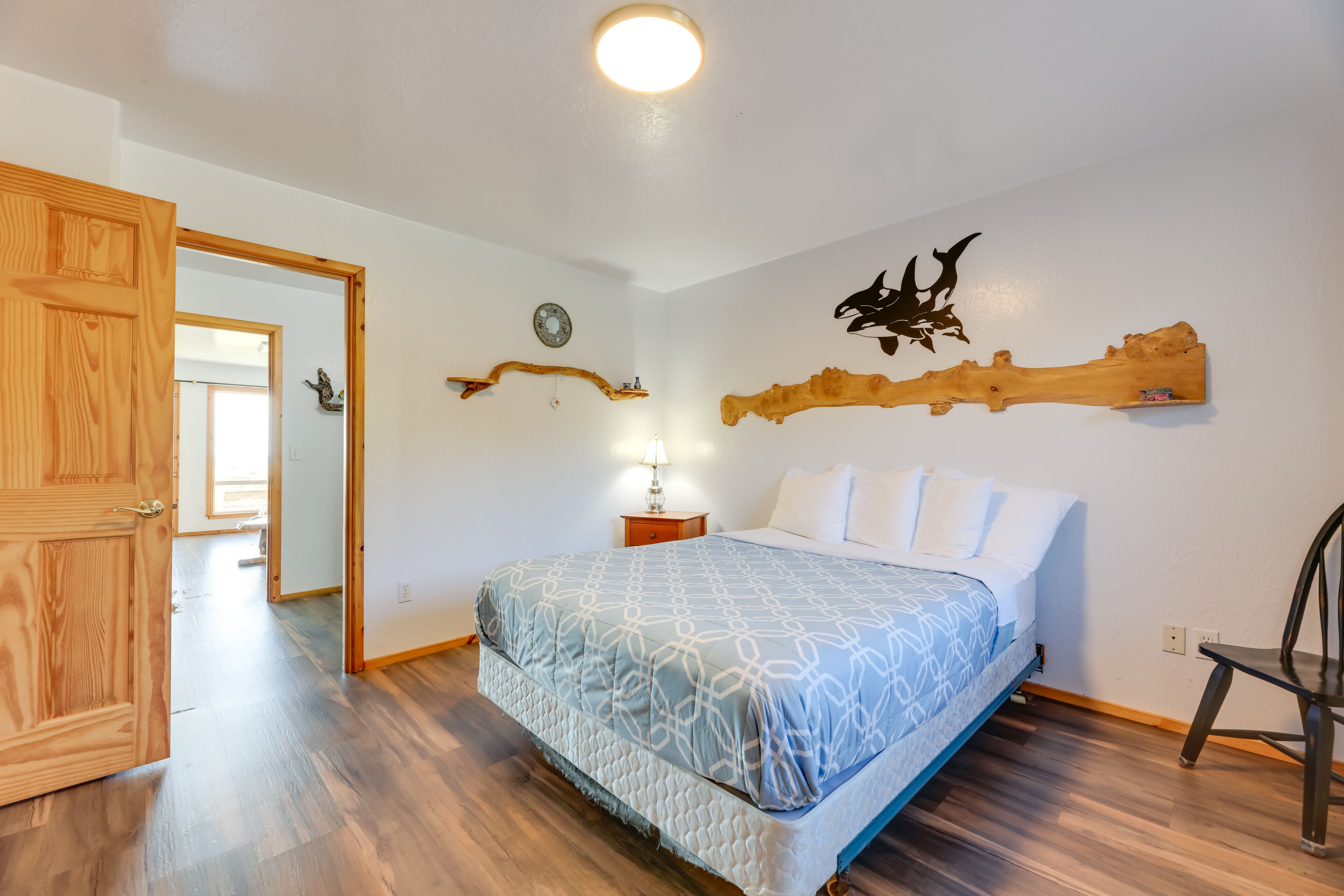 A bedroom with a bed, wooden floor, a nightstand with a lamp, a wall-mounted clock, and a decorative wooden piece.
