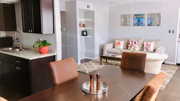 A modern kitchen with dark cabinets, a granite countertop, and a sink. There is a dining table with chairs and a decorative tray in the center. A living area with a sofa and cushions is visible in the background.