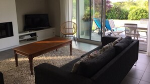 A modern living room with a sofa, a coffee table, a TV, and a large sliding glass door leading to an outdoor area with lounge chairs and a pool.