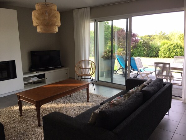 A modern living room with a sofa, a coffee table, a TV, and a large sliding glass door leading to an outdoor area with lounge chairs and a pool. - Villa du Ster, a stunning contemporary villa, within walking distance of Ster Beach (Penmarch)