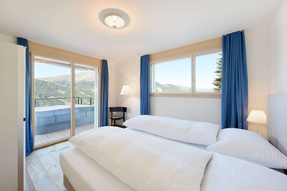 A hotel room with a bed, two bedside lamps, a chair, and a view of mountains through large windows.