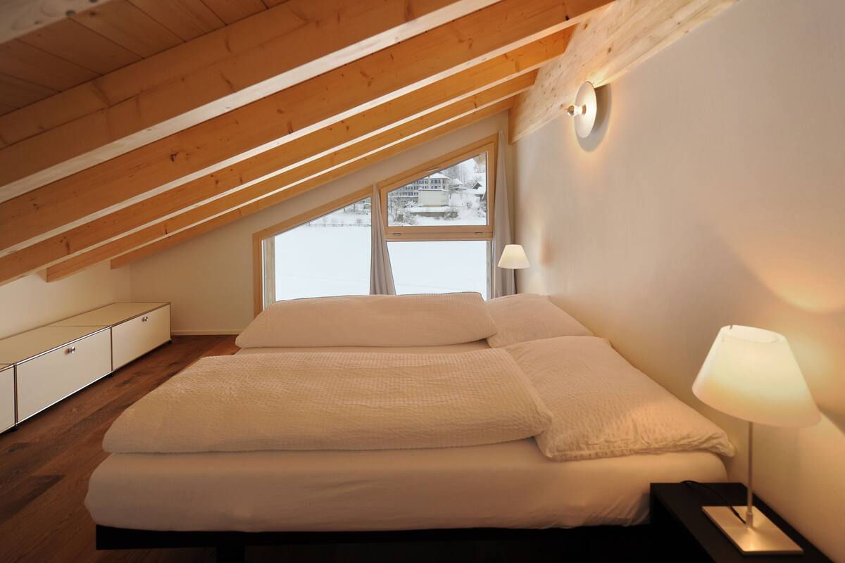 A bedroom with a sloped ceiling, a large bed, a bedside lamp, and a view of a snowy landscape through a window.