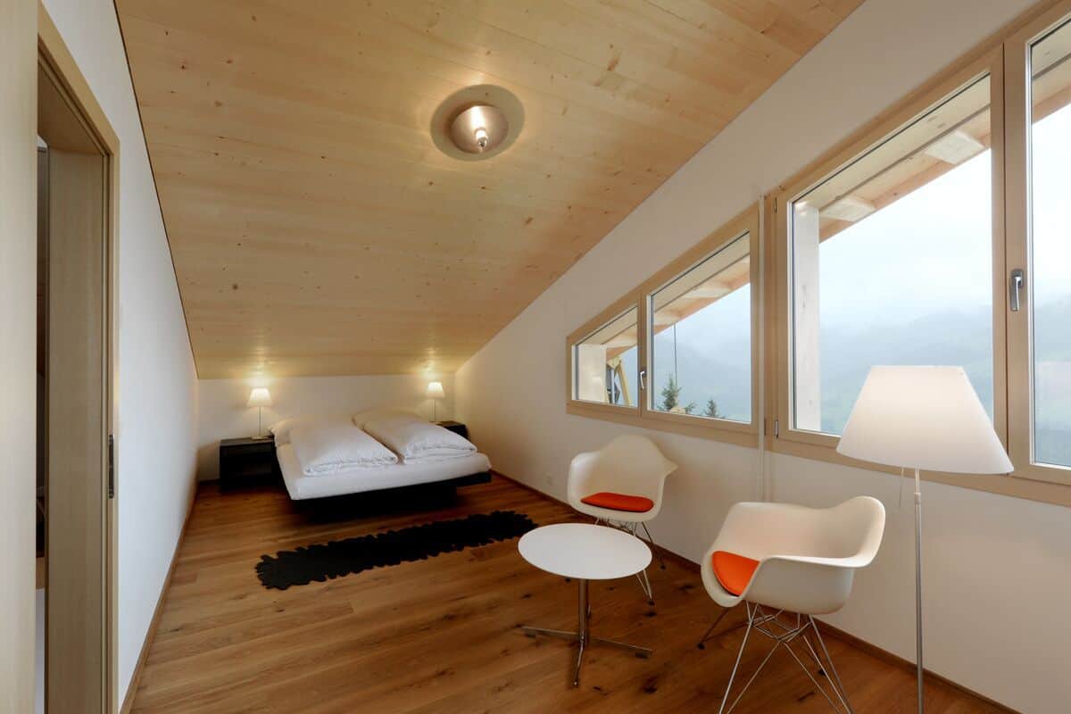 A modern bedroom with a wooden ceiling, a bed, a small table, two chairs, and a large window offering a view.