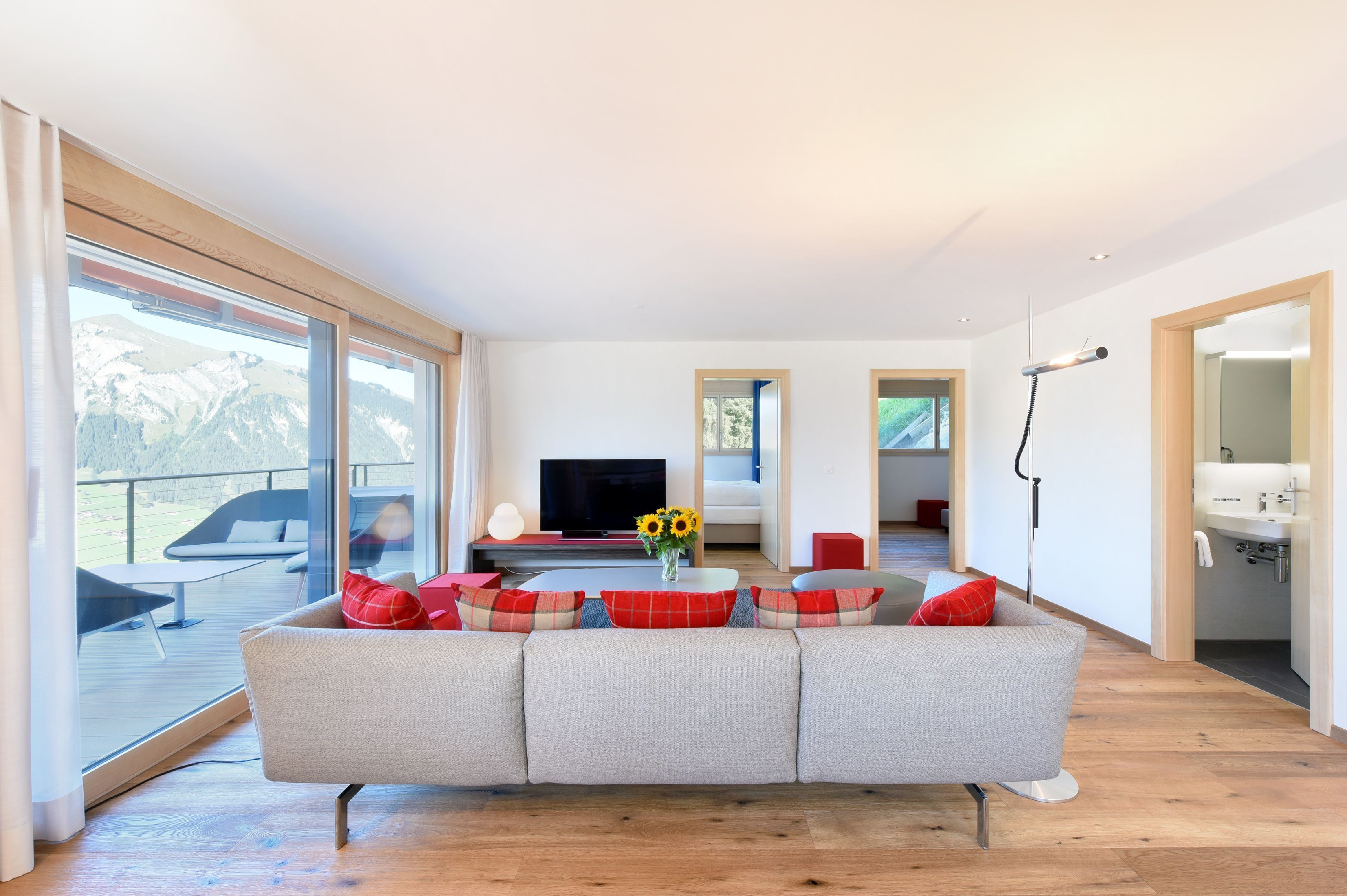 Een moderne woonkamer met een grijze bank, een flatscreen-tv en een groot raam met uitzicht op een terras.