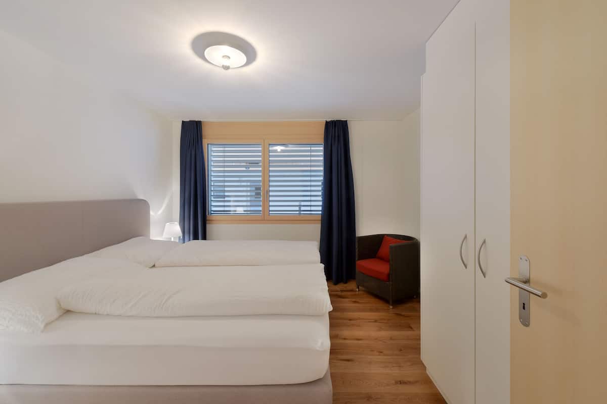 A bedroom with a bed, a chair, a wardrobe, and a window with curtains.