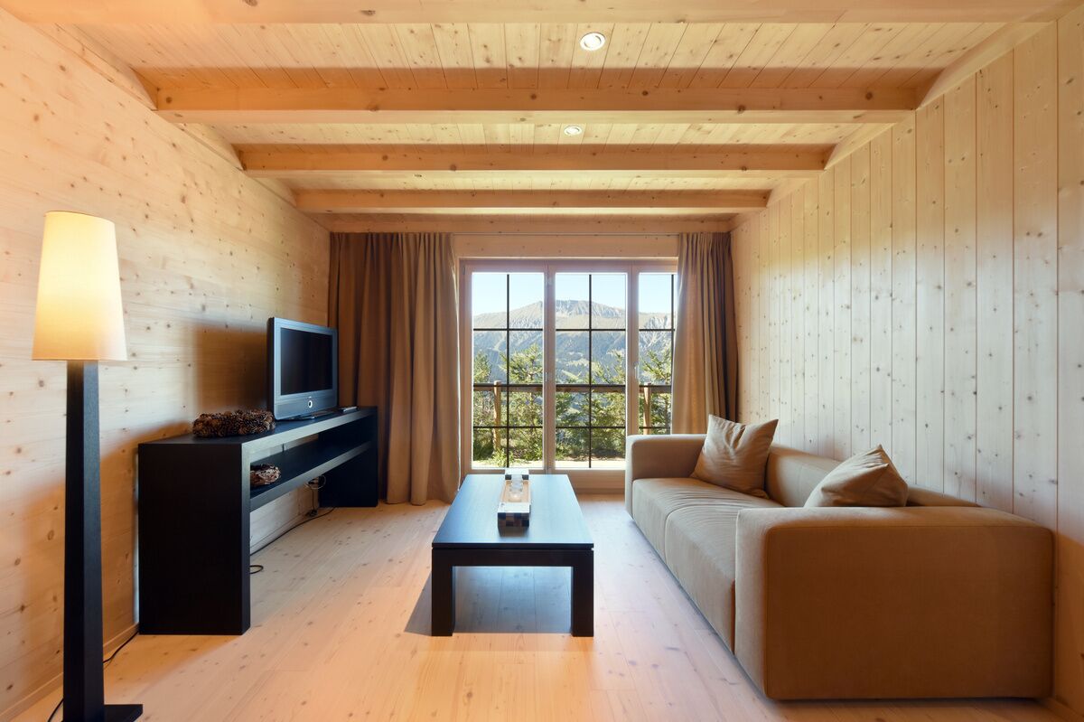 A cozy living room with a television, sofa, and coffee table, featuring wooden walls and a large window with a mountain view.