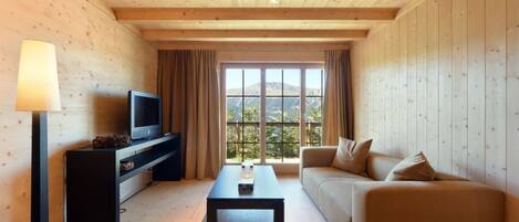 A cozy living room with a television, sofa, and coffee table, featuring wooden walls and a large window with a mountain view.