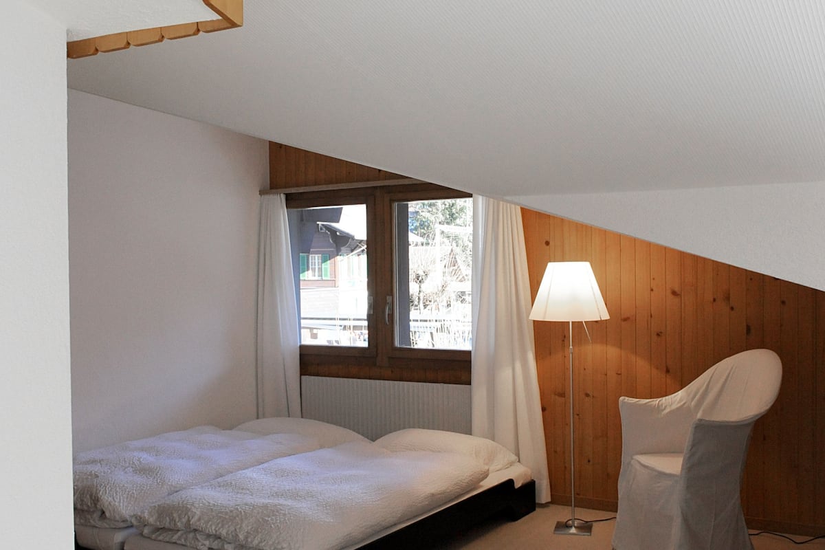 A bedroom with a single bed, a chair, a lamp, and a window with curtains.