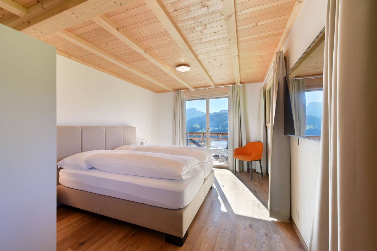 A bedroom with a wooden ceiling, a bed with white linens, a window with curtains, and a view of mountains.