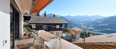 A balcony with a wooden table and chairs, overlooking a mountainous landscape.