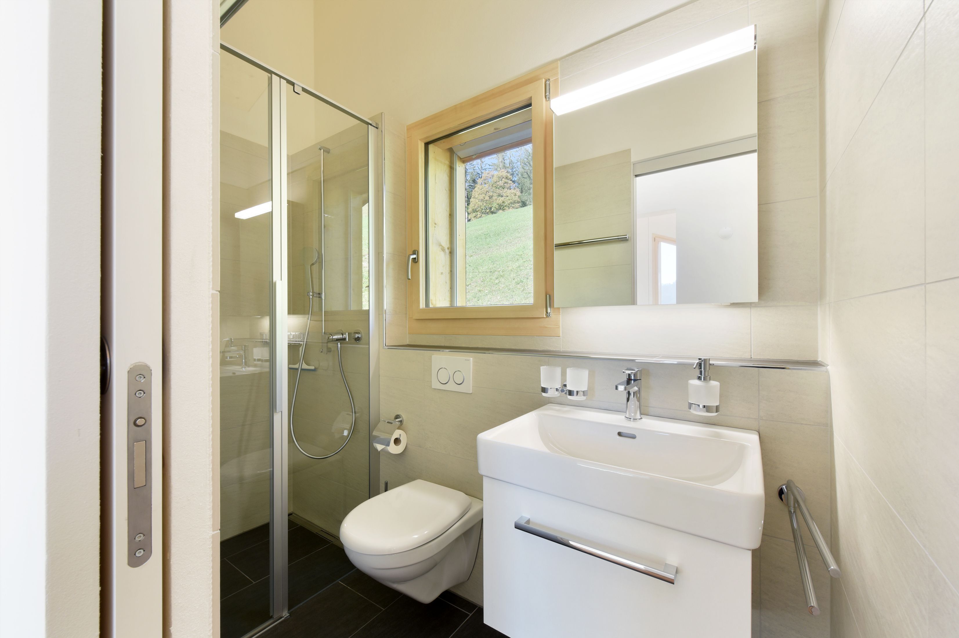A modern bathroom with a glass shower enclosure, a toilet, a sink with a mirror above it, and a window providing natural light.