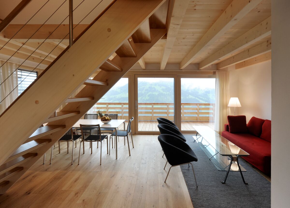 A modern interior with a wooden staircase, a dining area, a red sofa, and large windows offering a mountain view.