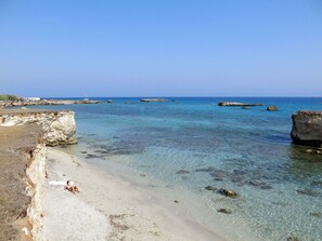 Une plage avec une eau claire, des affleurements rocheux et une personne allongée sur le sable.