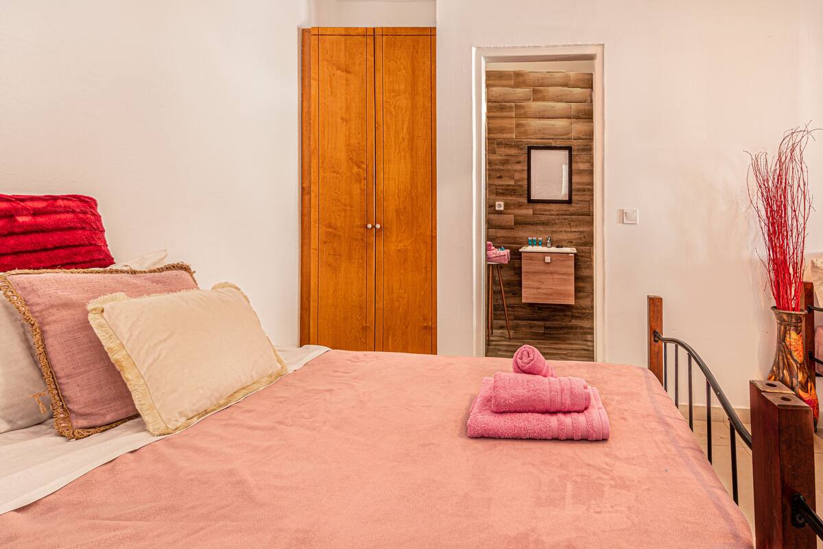 A bedroom with a bed, pillows, a wooden wardrobe, a bathroom with a sink, and a vase with red branches.