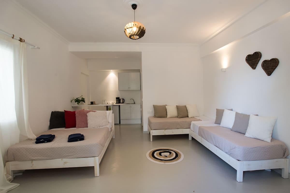 A modern, minimalist bedroom with two beds, a sofa, and a kitchen in the background.