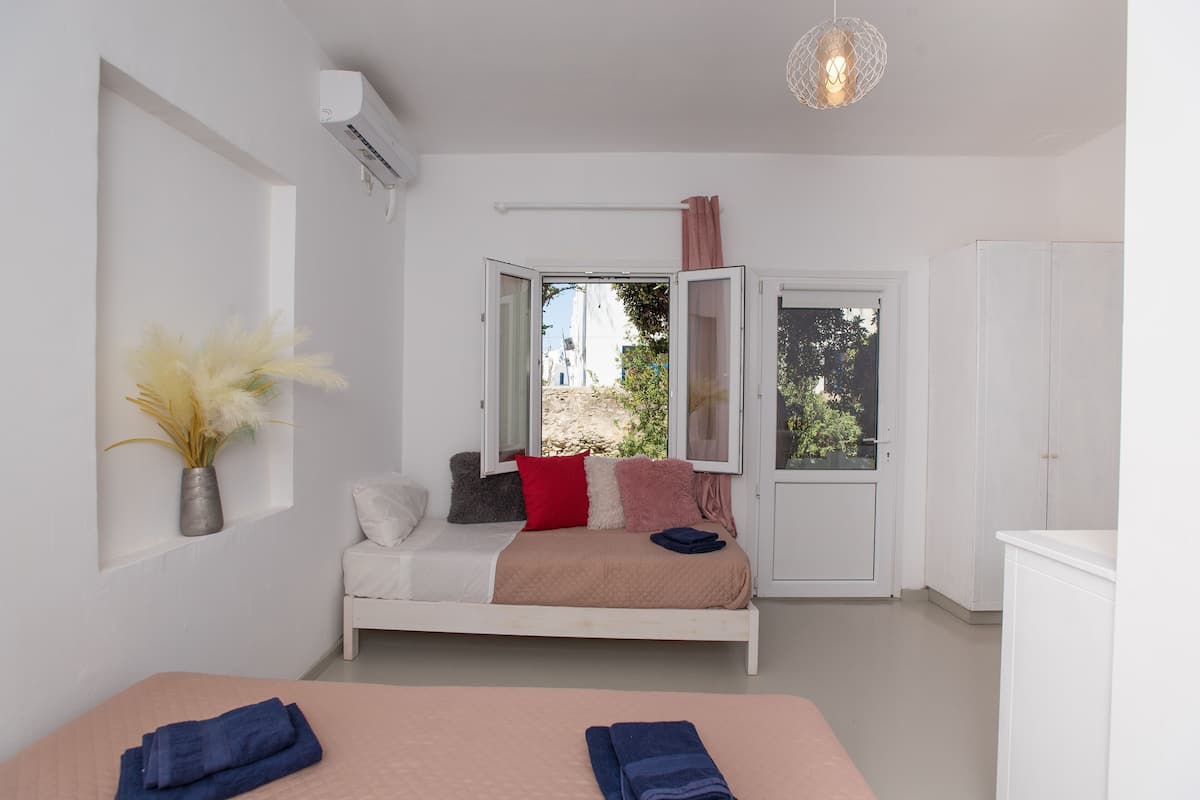 A bedroom with a bed, a bedside table, a window with curtains, a wall-mounted air conditioner, and a view of the outside.