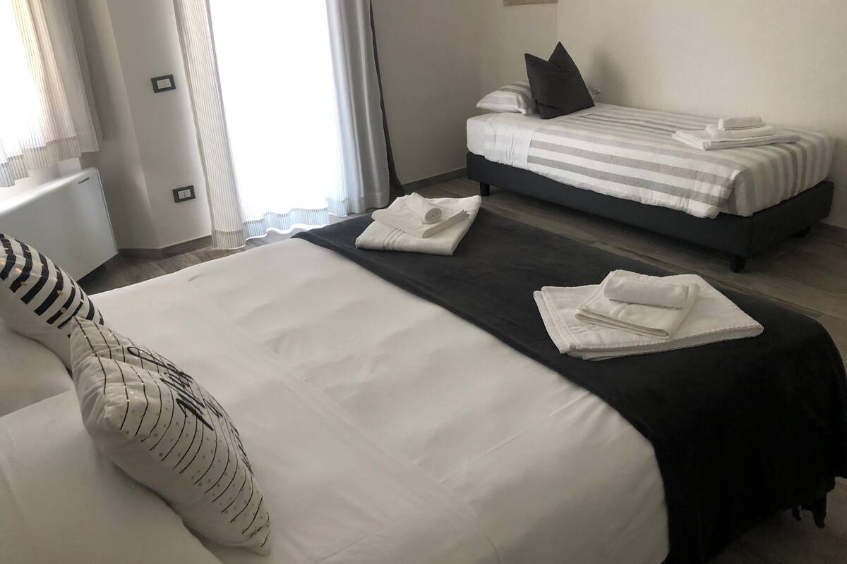 A hotel room with two beds, a large bed with a striped pillow and a smaller bed with a dark pillow. There are towels on the beds and a window with curtains.