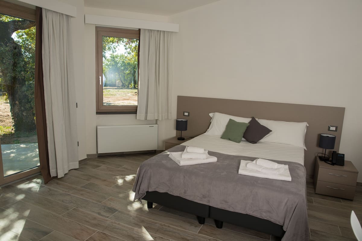 A bedroom with a large bed, a window with curtains, a bedside table with a lamp, and a radiator.