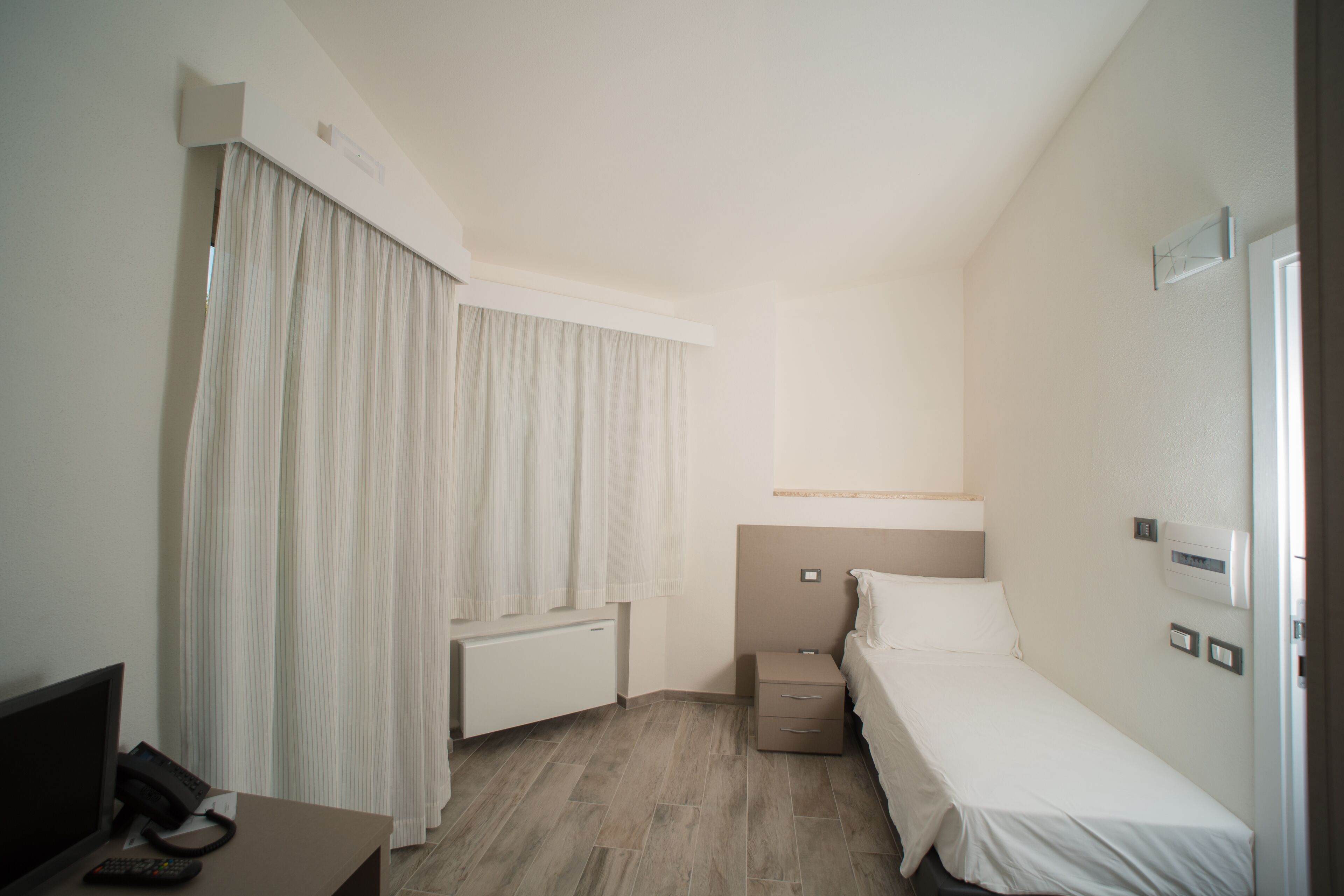 A hotel room with a bed, a bedside table, a desk with a telephone, and a window with curtains.
