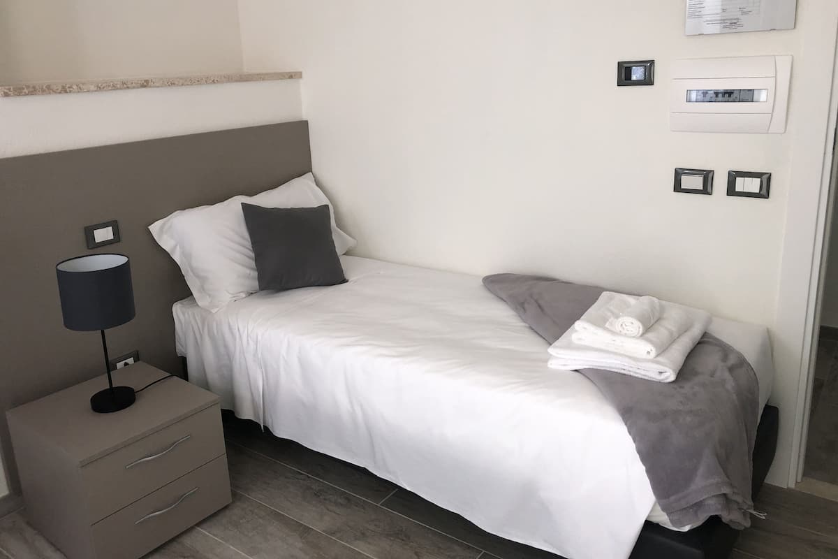 A neatly made bed with white bedding and a gray throw blanket, a bedside table with a lamp, and a wall-mounted thermostat.