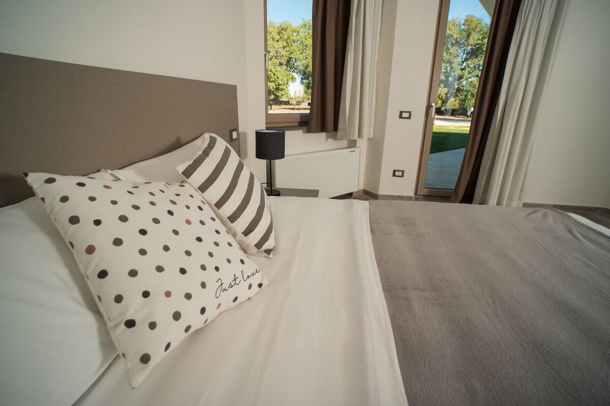 A neatly made bed with a polka dot pillow and striped pillow, a bedside lamp, and a view of a garden through an open door.